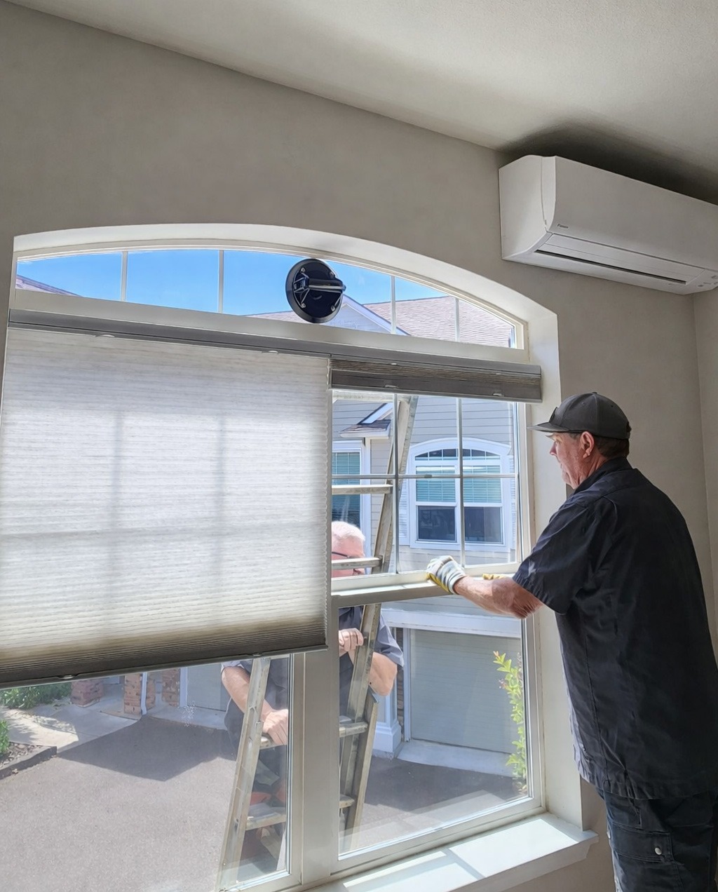 A worker adjusts a window treatment while another stands on a ladder outside the home. The scene highlights window installation or maintenance.