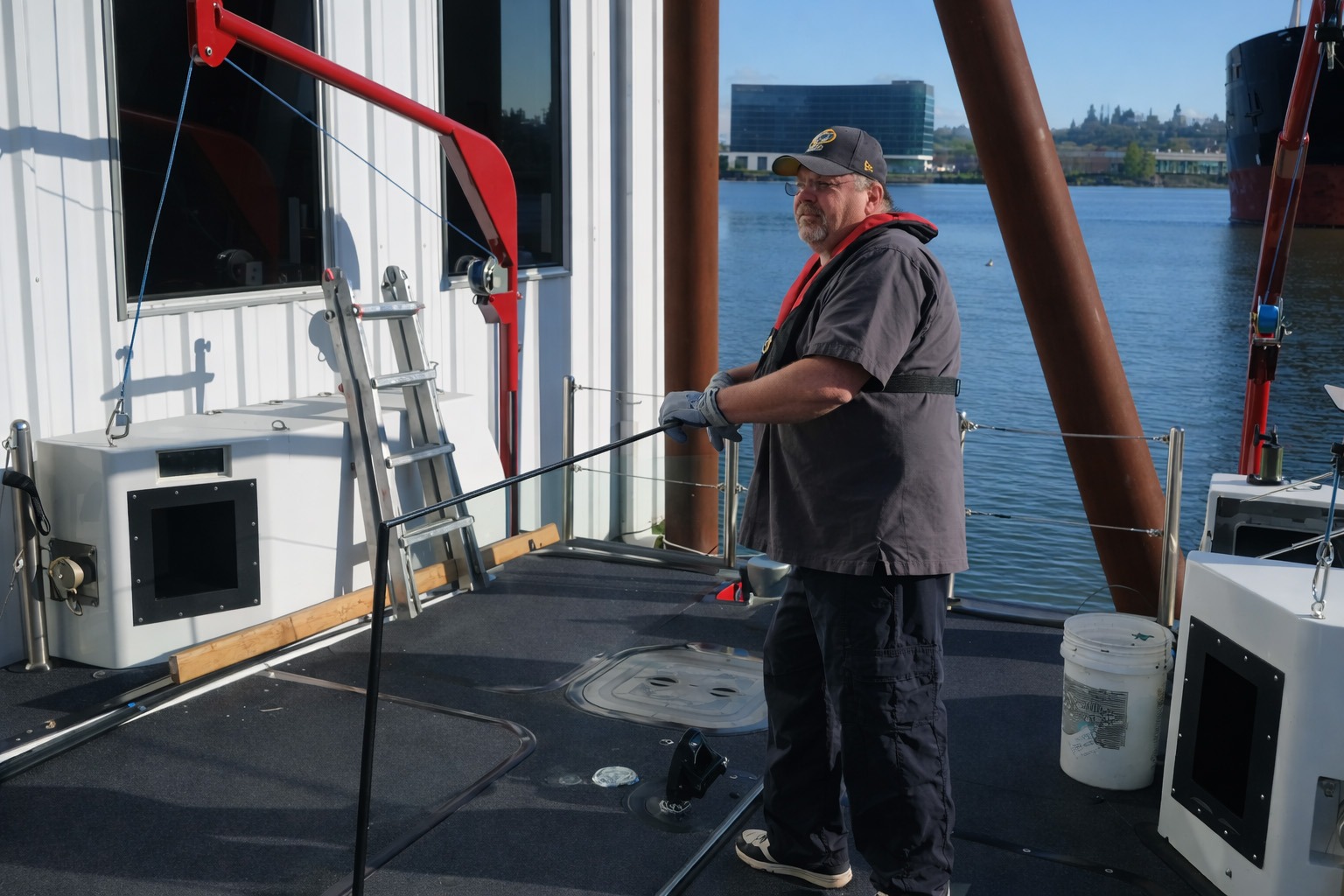 A man in a life jacket operates a boat's equipment on a sunny day, with a ladder and nearby water in the background.