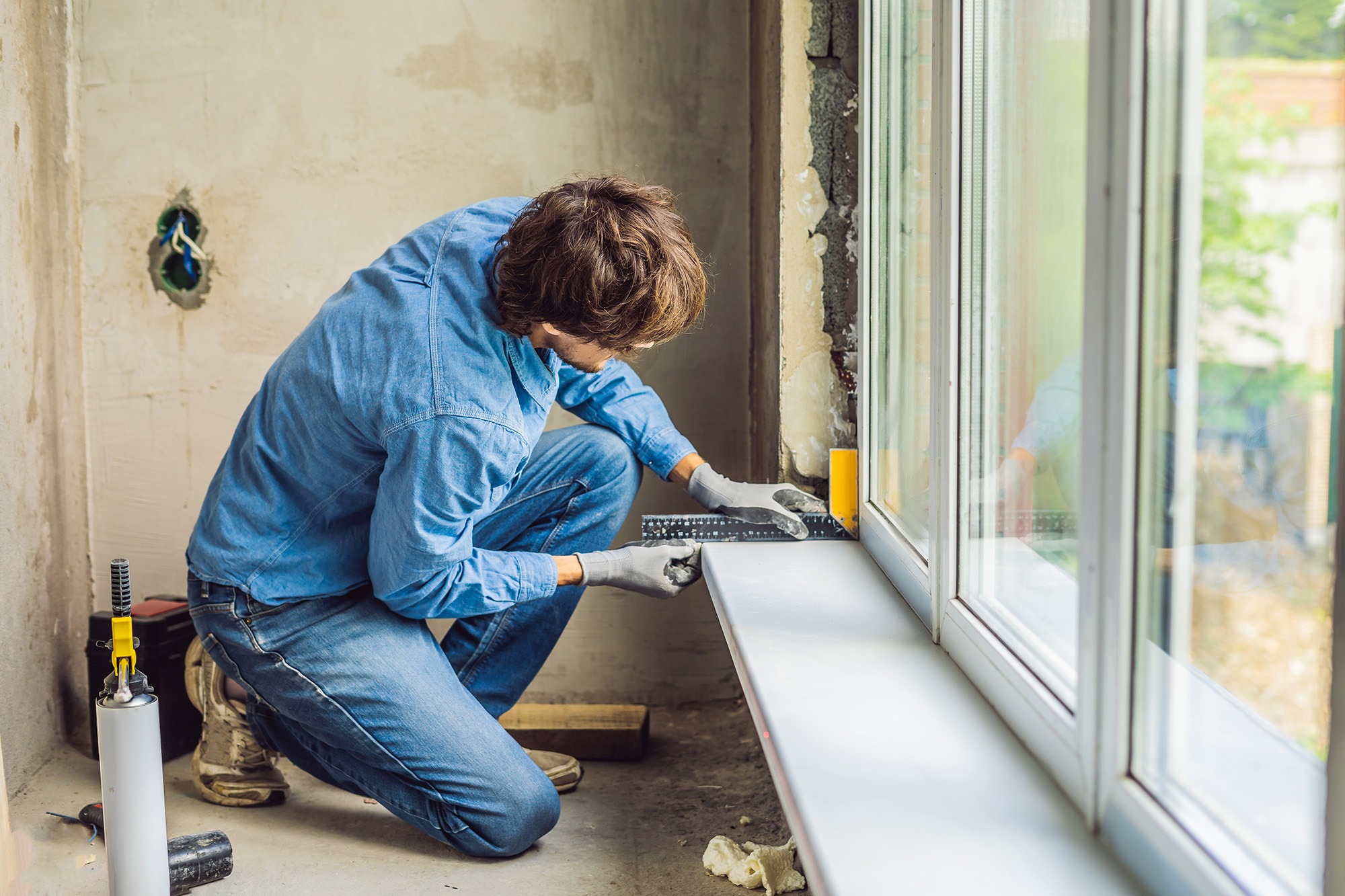 A worker installs a window sill in a partially renovated room, using a level tool to ensure proper alignment. Tools are visible nearby.
