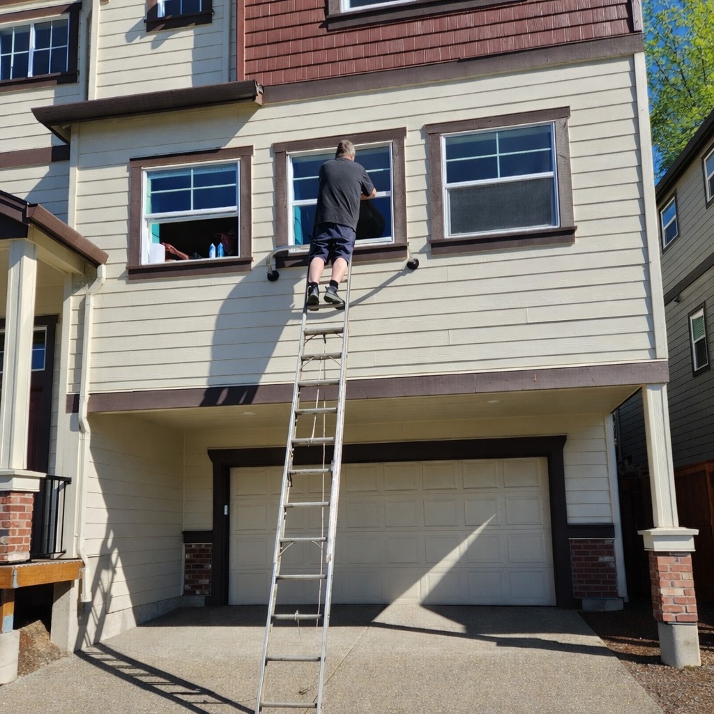 A person stands on a ladder cleaning the second-floor windows of a house, showcasing the home maintenance process.