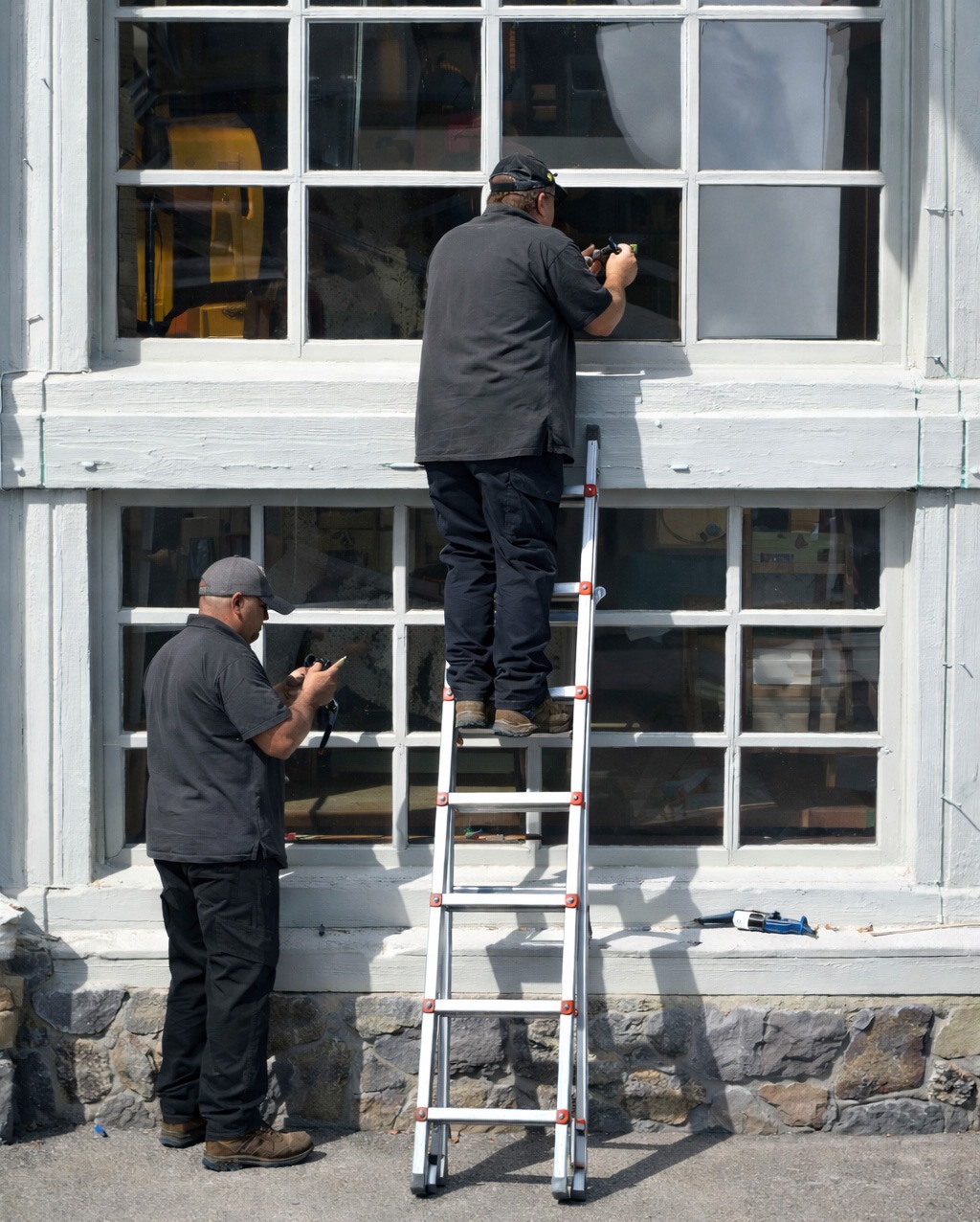 Two workers repairing windows: one on a ladder fixing the upper pane, another below holding tools. The setting features a stone wall backdrop.