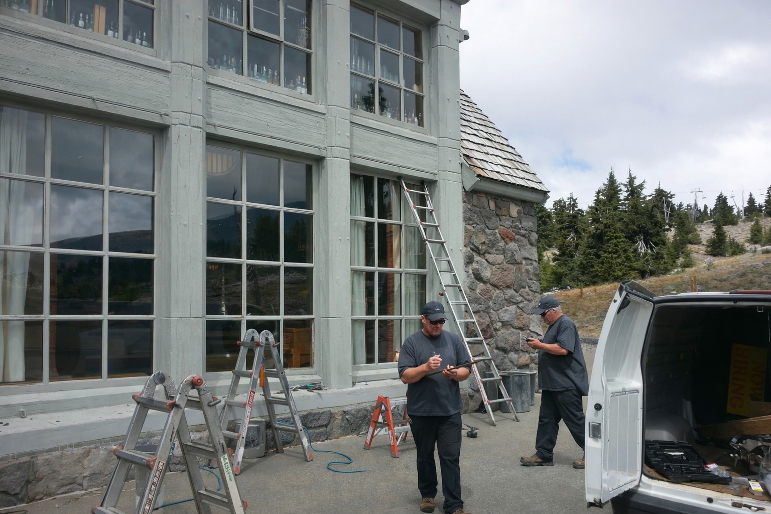 Two workers are performing maintenance on a large, windowed building exterior, using ladders and tools, with forested mountains in the background.