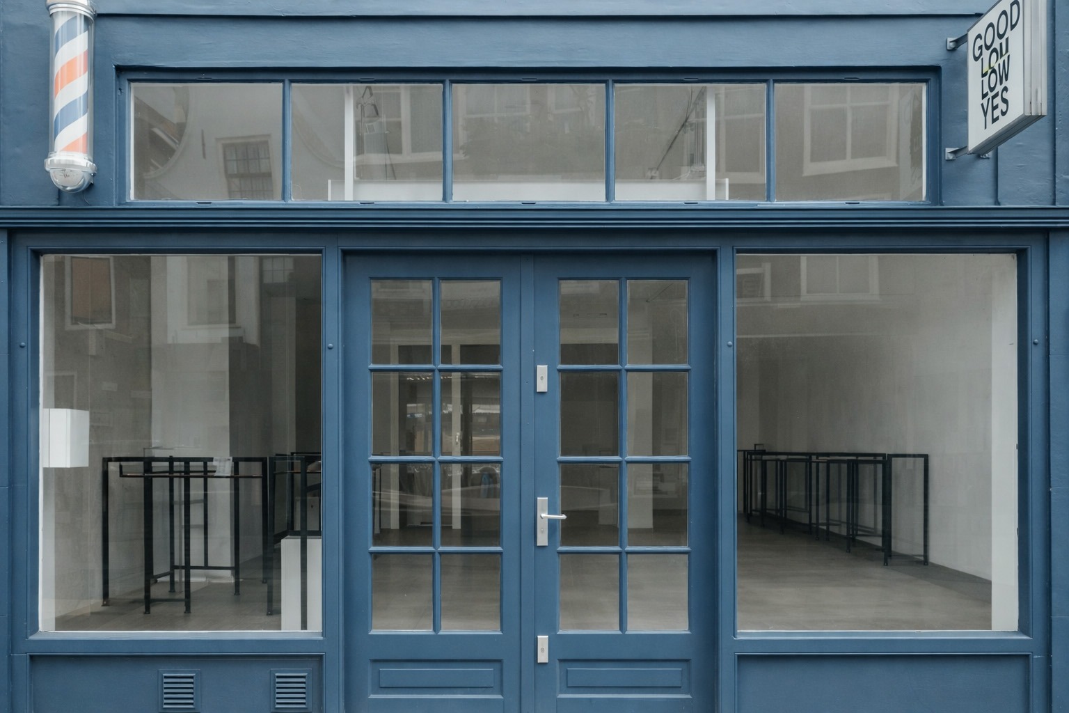 Blue storefront with a glass door and window displays. An empty interior features black display stands, suggesting a retail or gallery space.