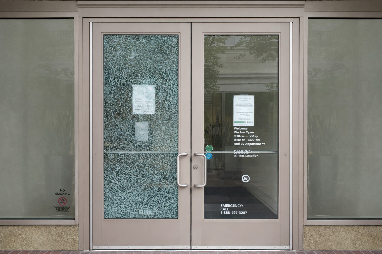 Shattered glass front door of a business with notices about operating hours and emergency contact information visible.