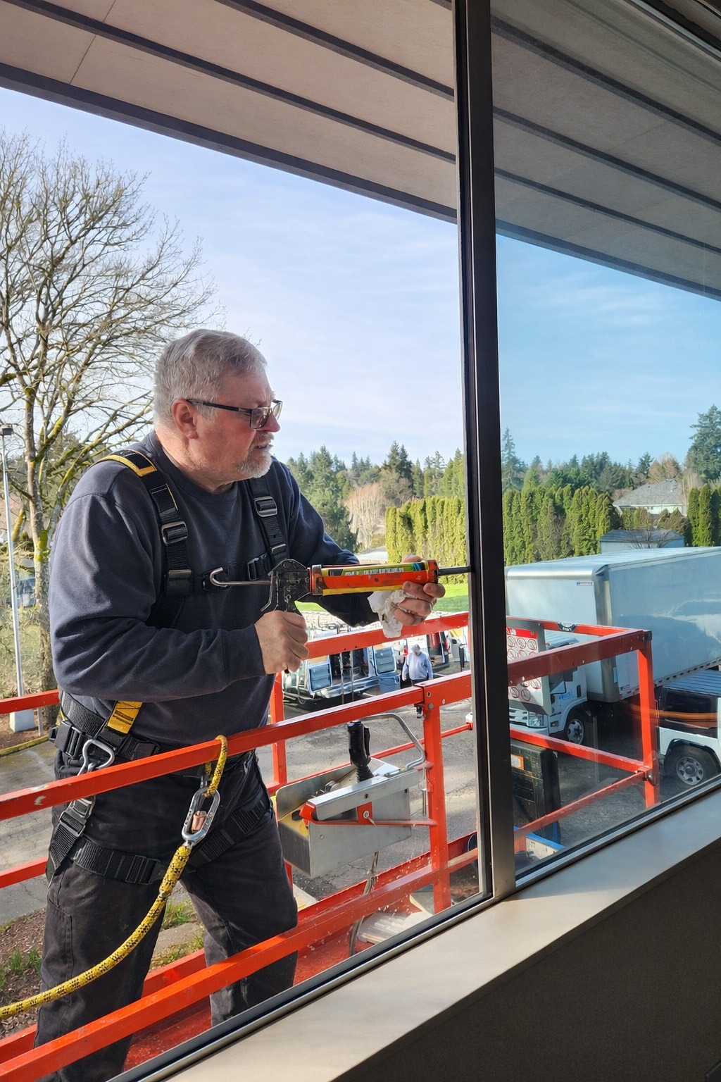 A worker in safety gear applies caulking to a window from a lift, with trucks and trees visible in the background, highlighting construction activity.