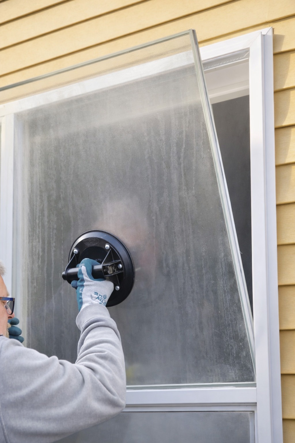 A person uses a suction cup to lift a fogged glass panel, demonstrating a window installation or repair process on a home.
