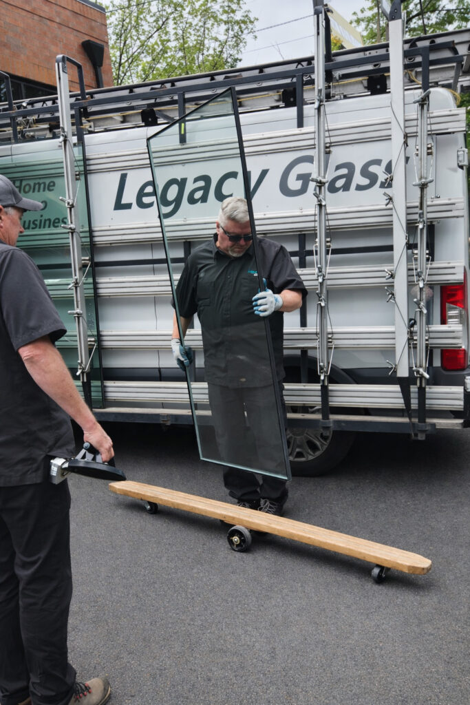 Technicians handle a large piece of glass using a vacuum lifter next to a van labeled "Legacy Glass." This image illustrates glass installation services.