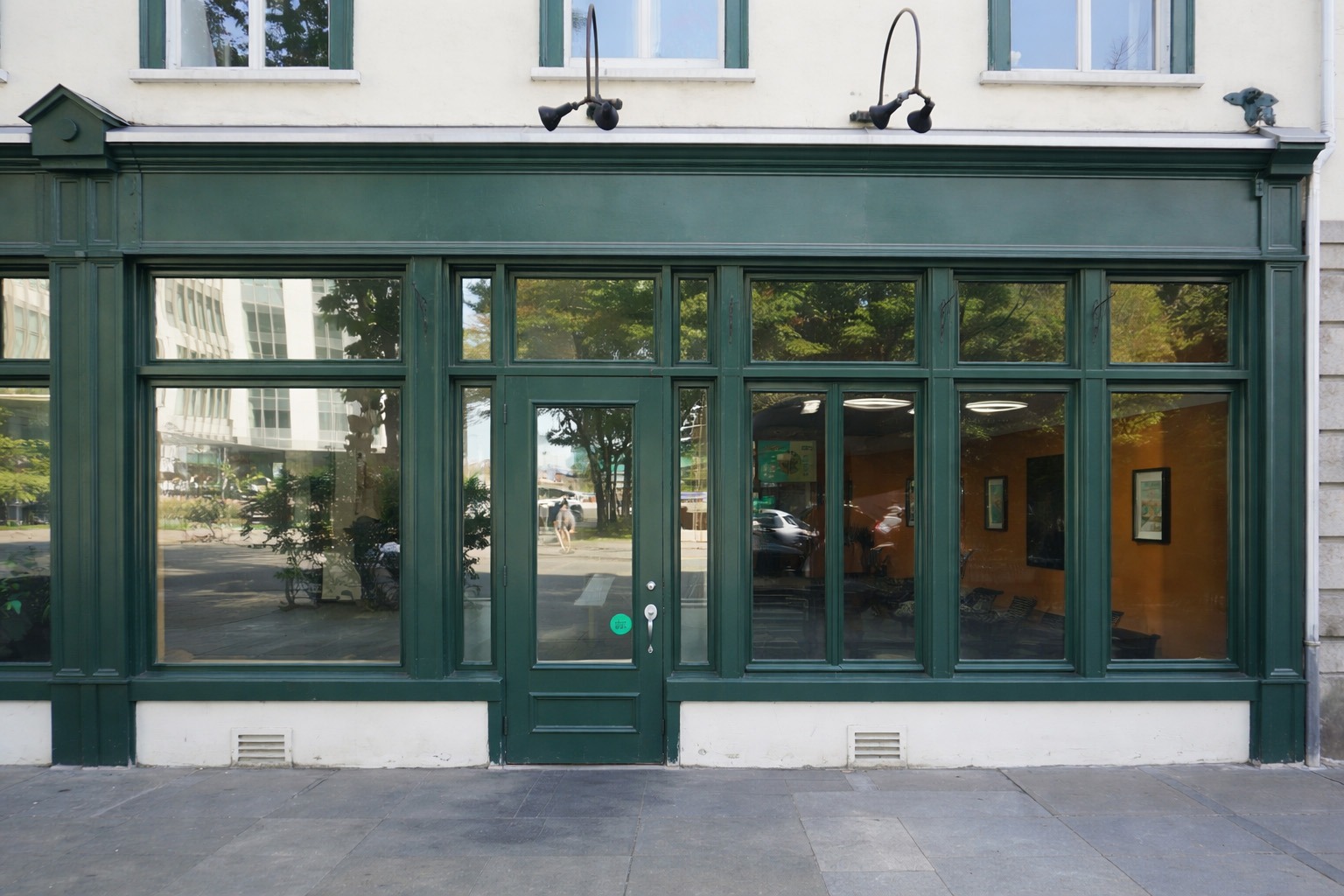 Green-framed storefront with large windows reflecting surrounding trees and buildings, suggesting a welcoming atmosphere for visitors.