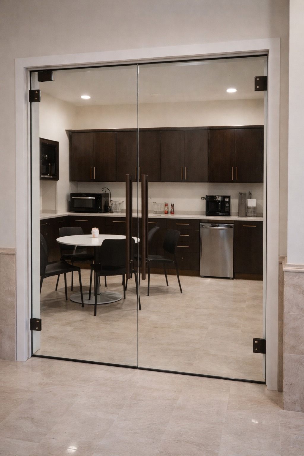 Modern kitchenette featuring dark wood cabinets, a round table with chairs, and appliances, enclosed by glass doors, ideal for communal dining.